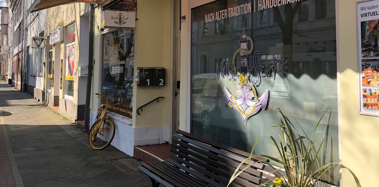Eine Außenansicht der Senfonie. man sieht die beiden Schaufenster im Halbschatten. Markisen sind ausgefahren. Ein gelbes Fahrrad und eine Parkbank steht vor dem Fenster.